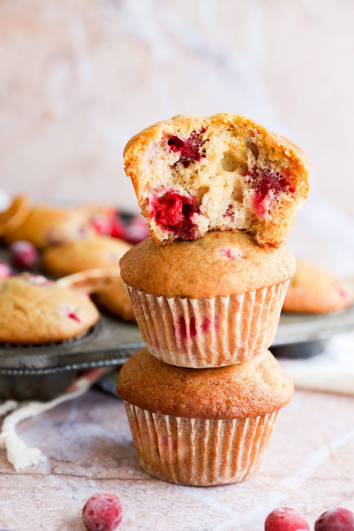 Two orange cranberry muffins are stacked on top of each other, with a third muffin—showing a bite taken out—resting on top. More muffins and scattered cranberries are in the background on a metal tray.