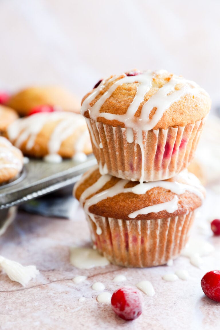 Two orange cranberry muffins stacked on top of each other, drizzled with white icing and studded with cranberries. More muffins and loose cranberries are scattered in the background on a light surface.