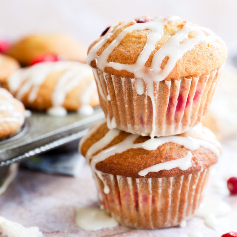 Two orange cranberry muffins stacked on top of each other, drizzled with white icing and studded with cranberries. More muffins and loose cranberries are scattered in the background on a light surface.