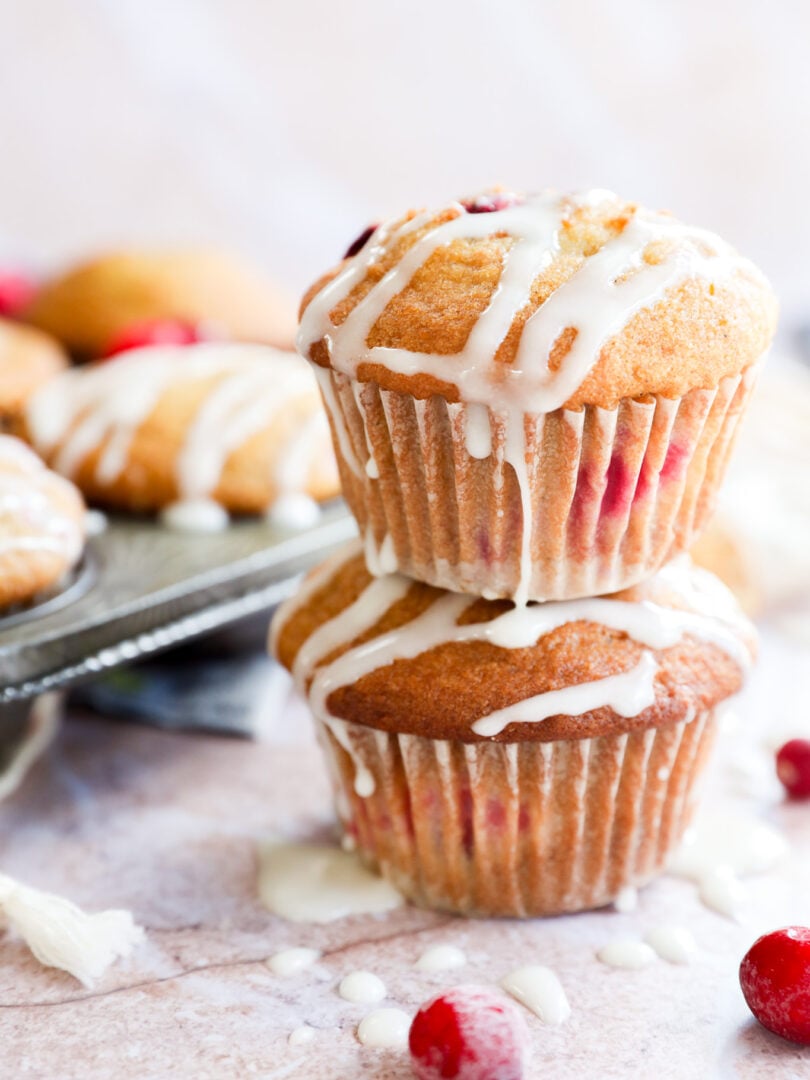 Two orange cranberry muffins stacked on top of each other, drizzled with white icing and studded with cranberries. More muffins and loose cranberries are scattered in the background on a light surface.