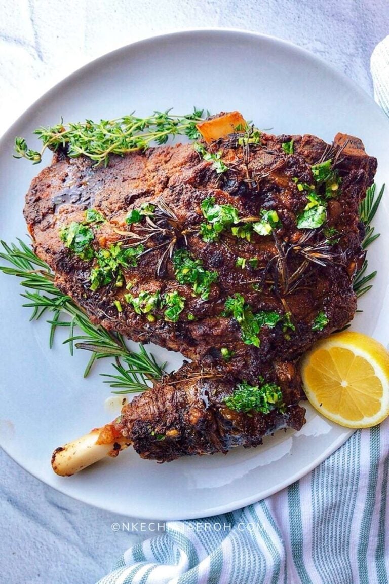 A plate with a large, cooked lamb shank garnished with chopped herbs and surrounded by fresh rosemary, thyme, and a lemon slice on the side. The meat looks juicy and seasoned.