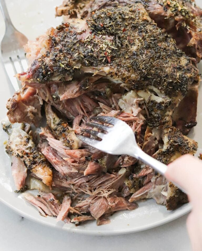 A close-up of a fork shredding tender, herb-crusted cooked lamb on a white plate. The meat appears juicy and well-seasoned, with visible herbs and spices on the surface.