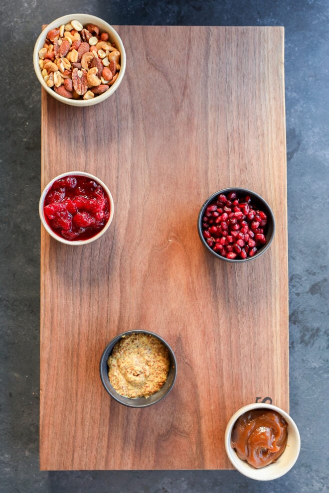 Five small bowls filled with mixed nuts, cranberry sauce, pomegranate seeds, whole grain mustard, and caramel sauce are arranged on a rectangular wooden board—perfect for a Thanksgiving charcuterie board—against a dark stone background.