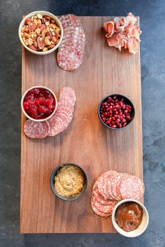 A Thanksgiving charcuterie board features a wooden platter with assorted salami, prosciutto, bowls of mixed nuts, pomegranate seeds, whole grain mustard, fruit preserves, and cranberry sauce, all arranged neatly on a dark surface.