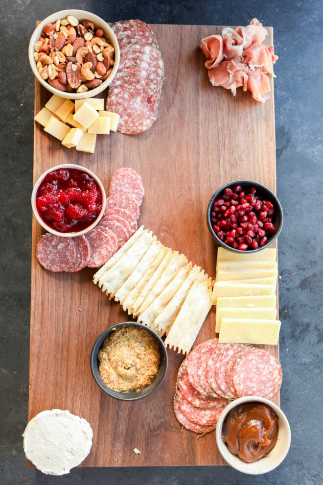 A Thanksgiving charcuterie board featuring assorted crackers, salami, cheeses, nuts, cranberry sauce, pomegranate seeds, prosciutto, mustard, goat cheese, and a small bowl of brown chutney arranged neatly on a wooden platter.