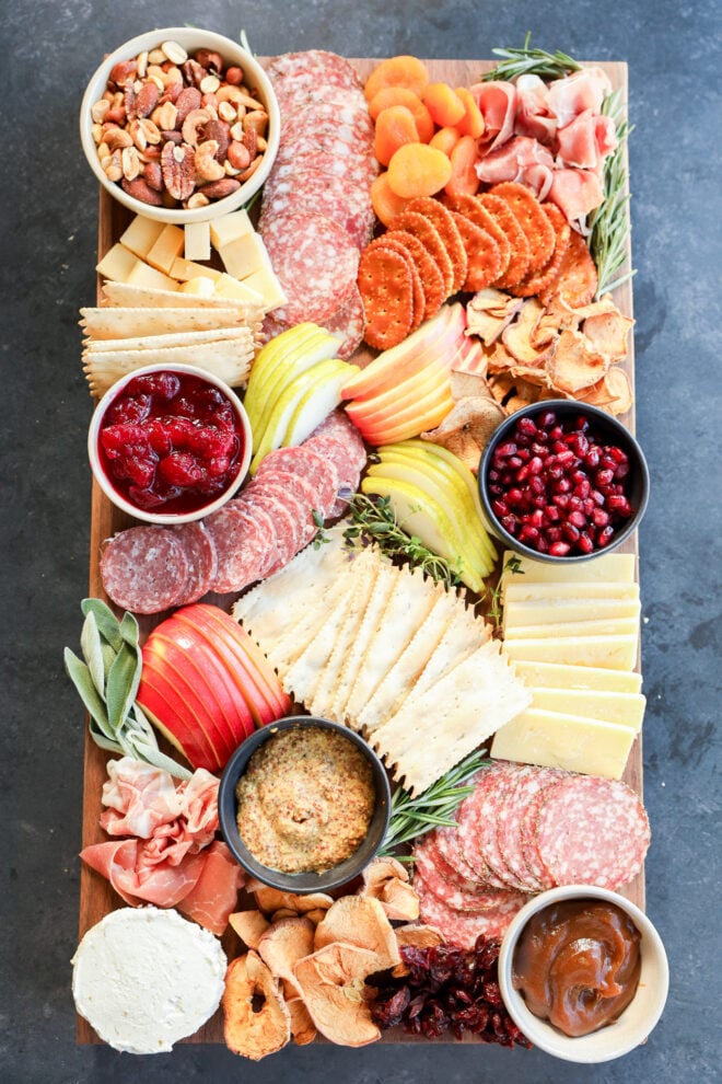 A rectangular wooden Thanksgiving charcuterie board topped with assorted cheeses, sliced meats, crackers, dried fruits, apple slices, nuts, bowls of mustard, cranberry sauce, pomegranate seeds, and caramel dip arranged artfully.