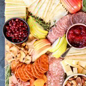 A Thanksgiving charcuterie board with salami, prosciutto, crackers, cheeses, apple and pear slices, dried apricots, nuts, pomegranate seeds, dried fruit chips, mustard, cranberry sauce, and herb garnishes.