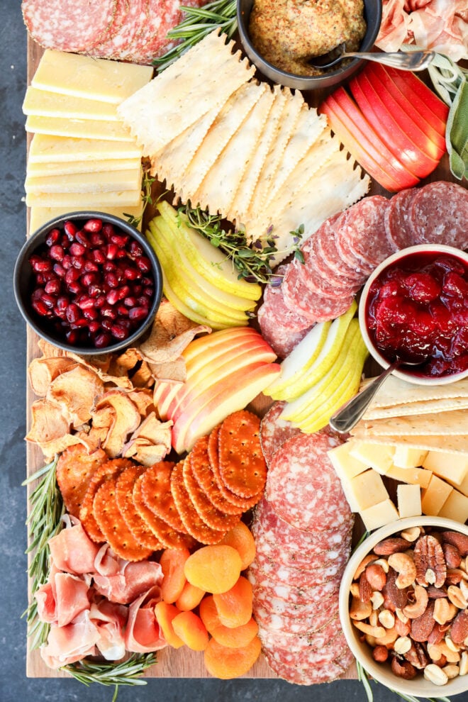 A Thanksgiving charcuterie board with salami, prosciutto, crackers, cheeses, apple and pear slices, dried apricots, nuts, pomegranate seeds, dried fruit chips, mustard, cranberry sauce, and herb garnishes.