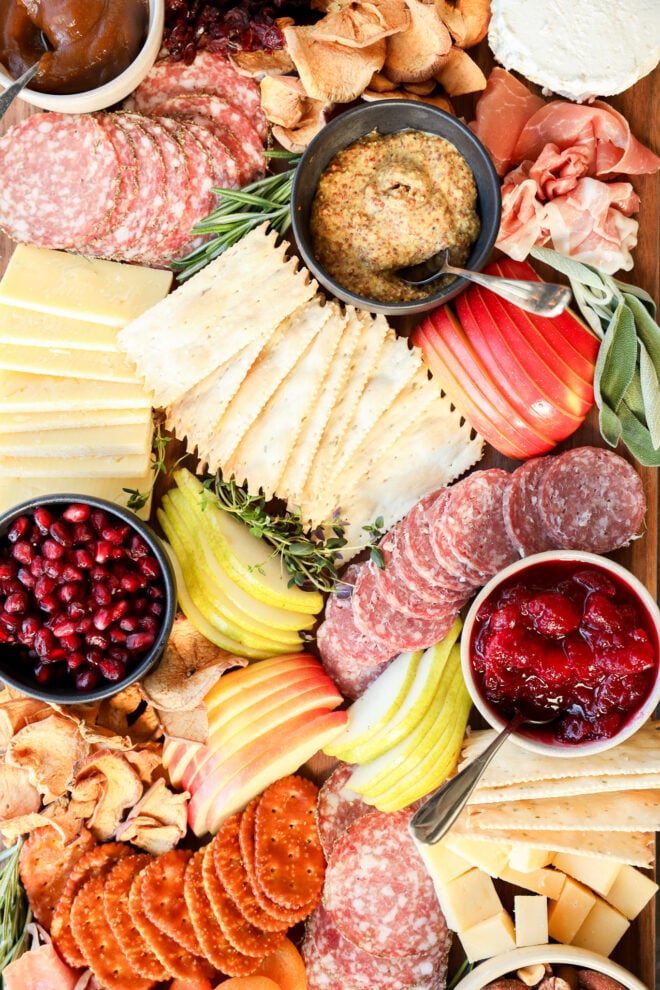 A colorful Thanksgiving charcuterie board with sliced meats, cheeses, crackers, apple and pear slices, pomegranate seeds, mustards, fruit preserves, chips, and herbs—all artfully arranged for your holiday gathering.