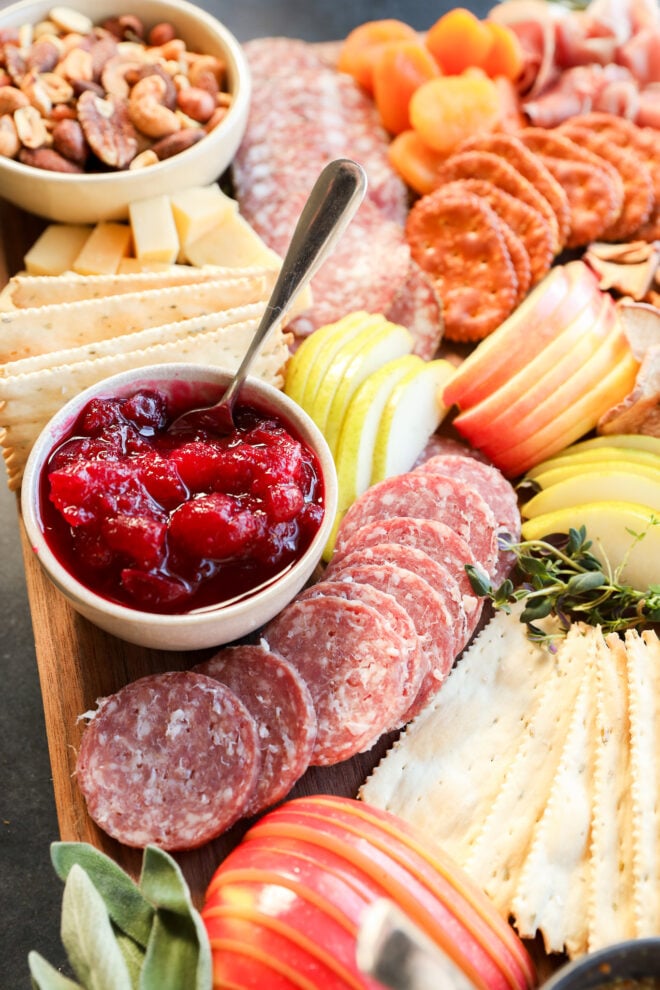 A Thanksgiving charcuterie board with sliced salami, apples, pears, crackers, dried apricots, mixed nuts, and a bowl of red fruit jam with a spoon, all arranged neatly on a wooden platter.