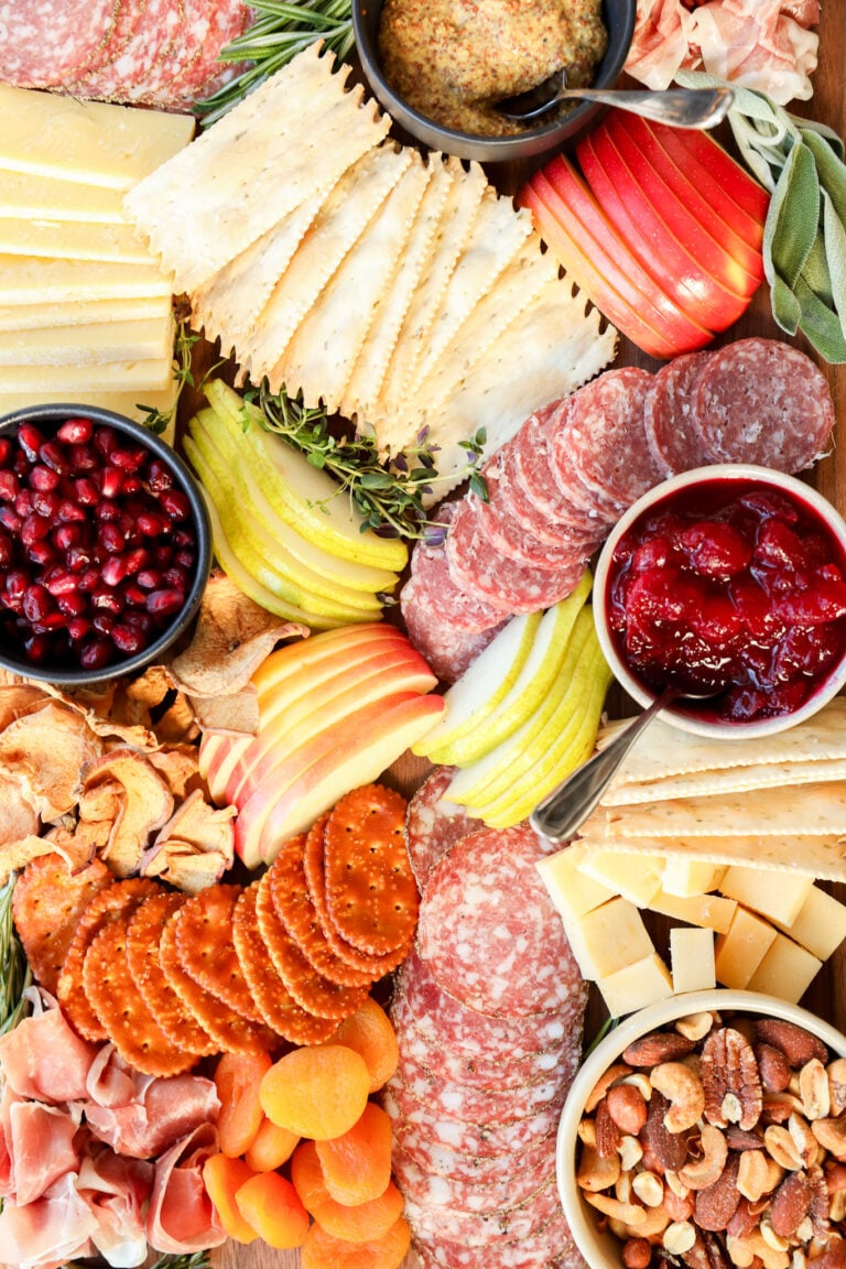 A colorful Thanksgiving charcuterie board with assorted cheeses, sliced salami, crackers, apple and pear slices, dried apricots, nuts, prosciutto, pomegranate seeds, grainy mustard, and a bowl of cranberry sauce.