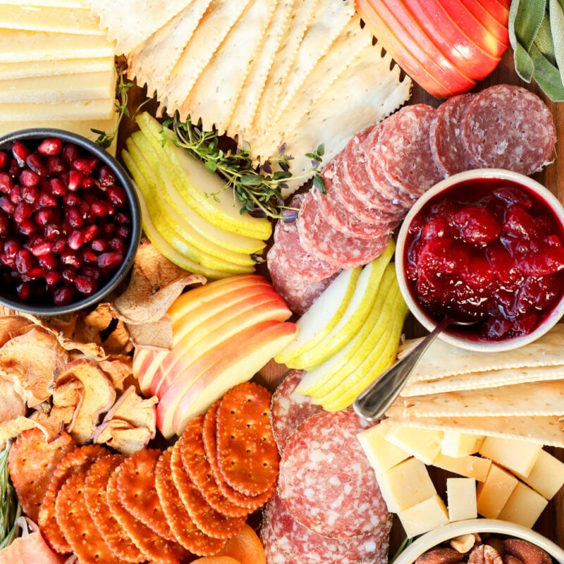 A colorful Thanksgiving charcuterie board with assorted cheeses, sliced salami, crackers, apple and pear slices, dried apricots, nuts, prosciutto, pomegranate seeds, grainy mustard, and a bowl of cranberry sauce.