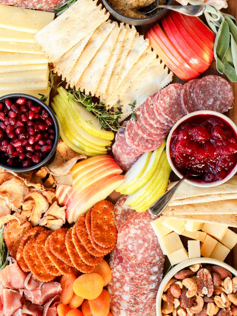 A colorful Thanksgiving charcuterie board with assorted cheeses, sliced salami, crackers, apple and pear slices, dried apricots, nuts, prosciutto, pomegranate seeds, grainy mustard, and a bowl of cranberry sauce.
