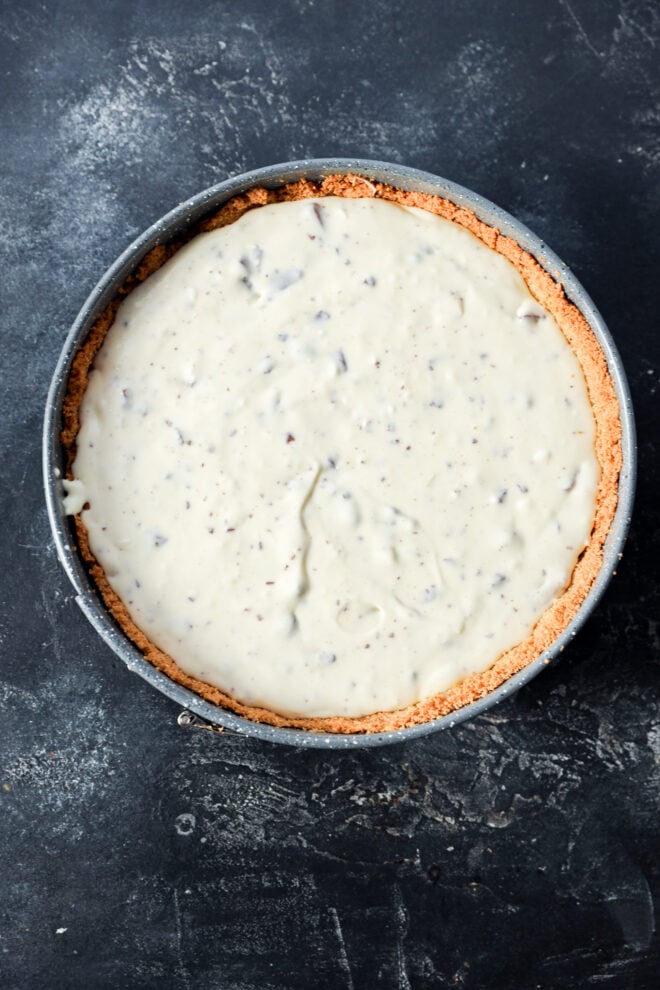 Before baking a round Toblerone cheesecake with a crumbly crust sits in a baking pan on a dark, textured surface. The creamy filling is smooth with small bits of chocolate mixed in.
