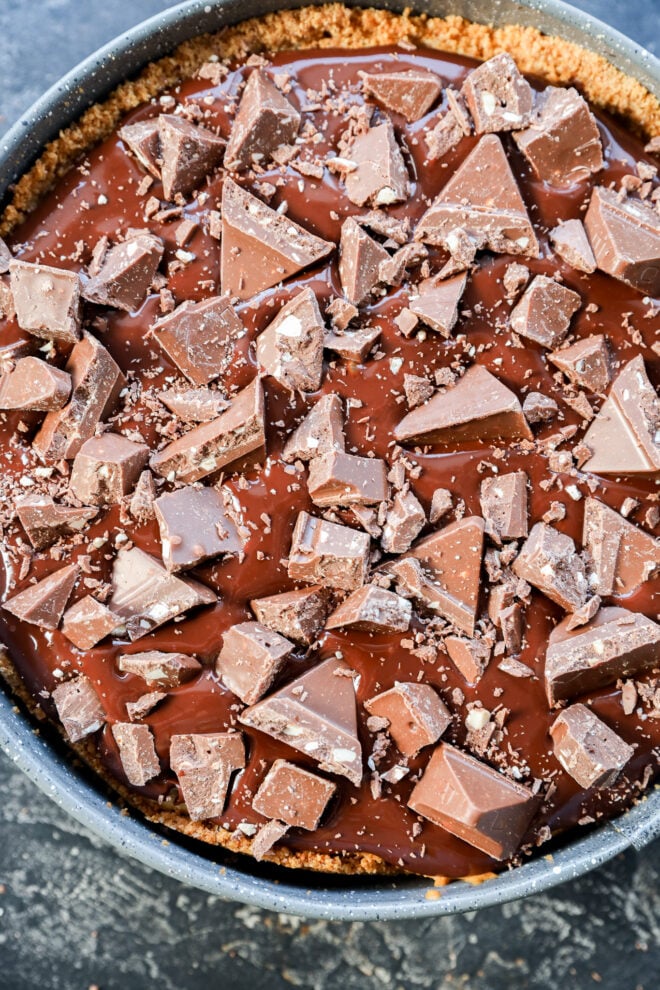 A close-up of a chocolate pie reminiscent of a toblerone cheesecake, topped with chunks of milk chocolate on a graham cracker crust, with smooth chocolate filling visible underneath the chocolate pieces.