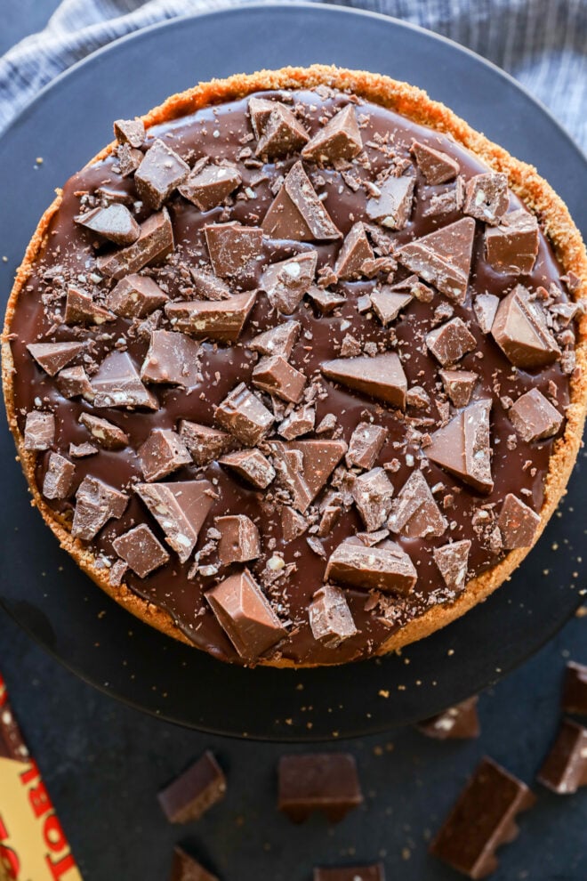 A round Toblerone cheesecake topped with a glossy chocolate layer and large chunks of milk chocolate, set on a dark surface with pieces of chocolate scattered around.