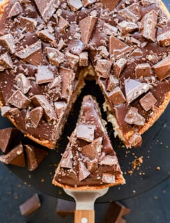 A Toblerone cheesecake topped with large chunks of chocolate, with one slice being lifted out on a metal spatula, revealing a creamy interior and scattered chocolate pieces on and around the cake.