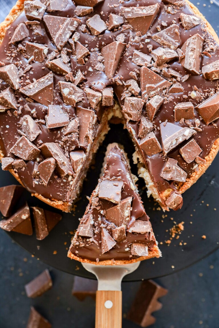 A Toblerone cheesecake topped with large chunks of chocolate, with one slice being lifted out on a metal spatula, revealing a creamy interior and scattered chocolate pieces on and around the cake.
