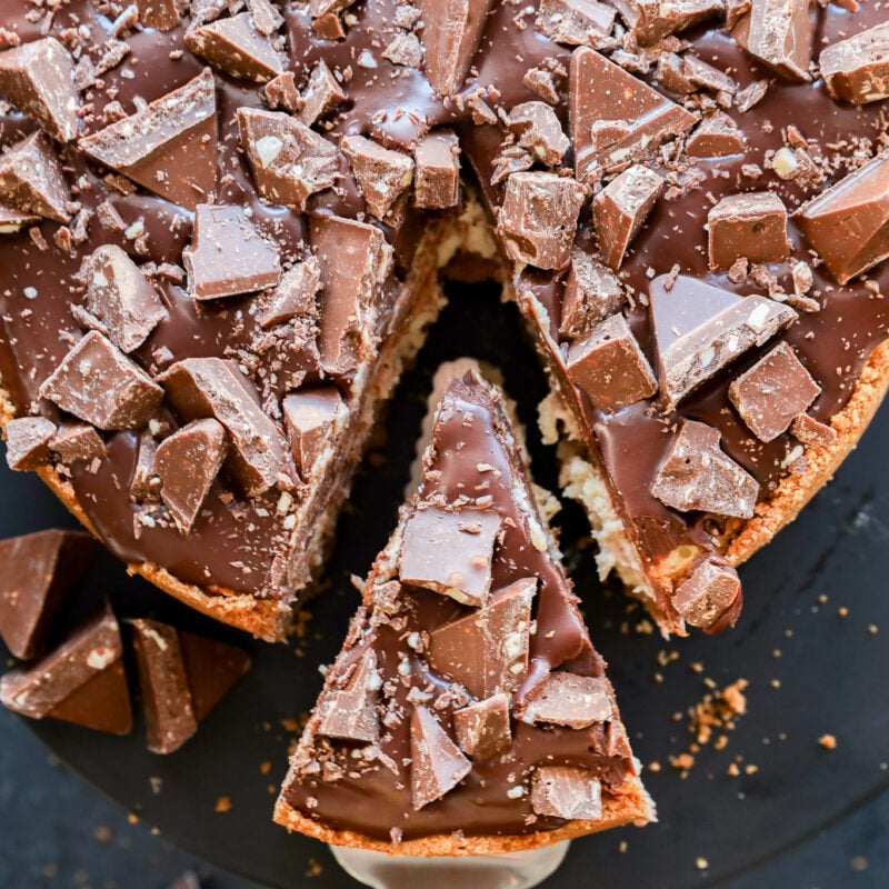 A Toblerone cheesecake topped with large chunks of chocolate, with one slice being lifted out on a metal spatula, revealing a creamy interior and scattered chocolate pieces on and around the cake.