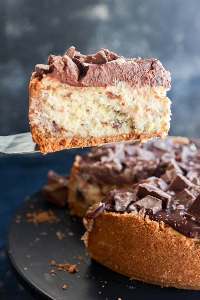 A close-up of a slice of Toblerone cheesecake with chocolate chunks on top, being lifted from a whole cheesecake on a black cake stand.