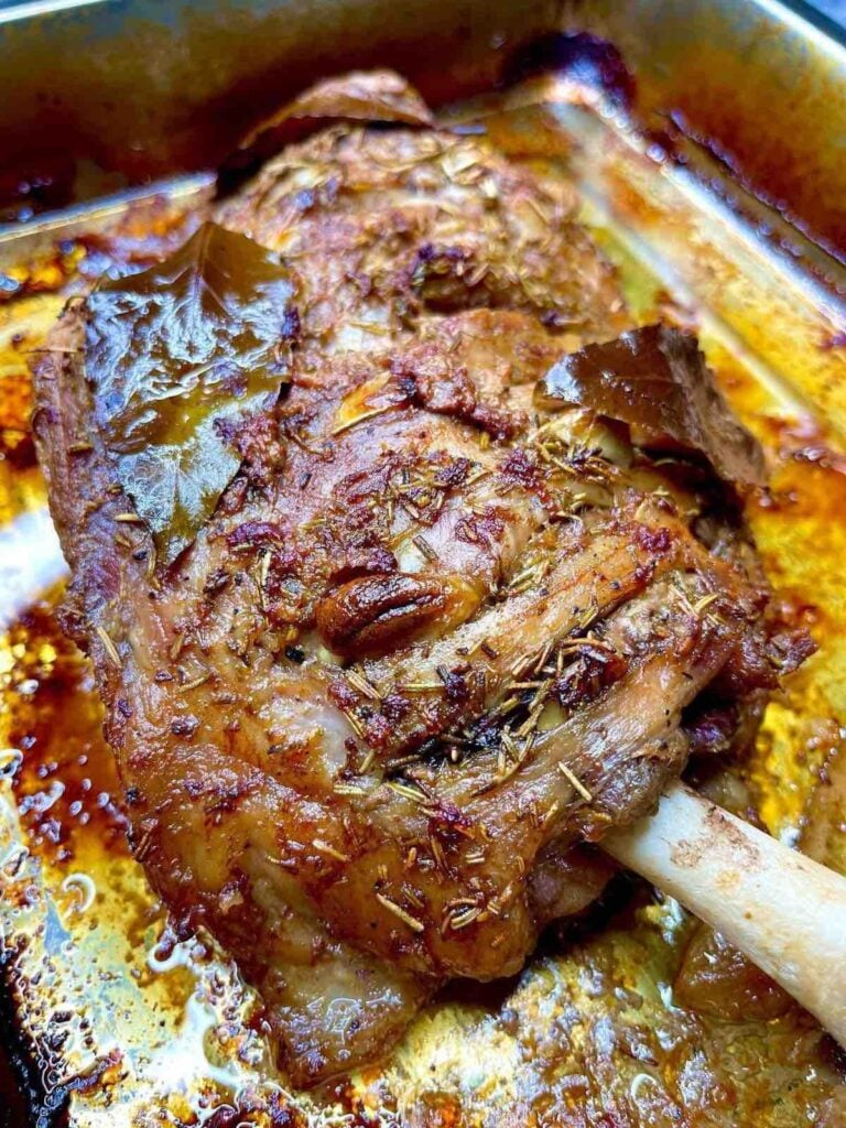 Close-up of a roasted lamb shank in a baking tray, seasoned with herbs and garnished with bay leaves, showing a crispy, golden-brown exterior and juices pooled around the meat.