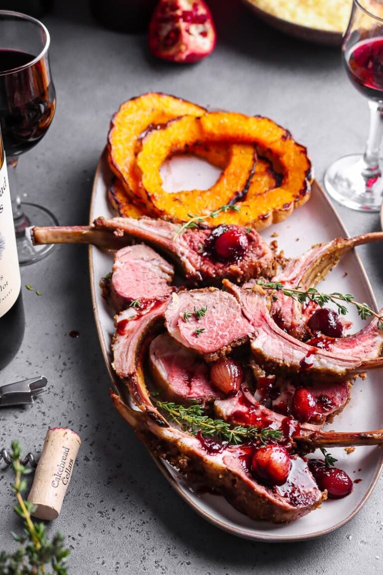 A platter with herb-crusted rack of lamb slices topped with red sauce and herbs, served with roasted squash rings. Two glasses of red wine and a wine bottle are nearby on a gray table.