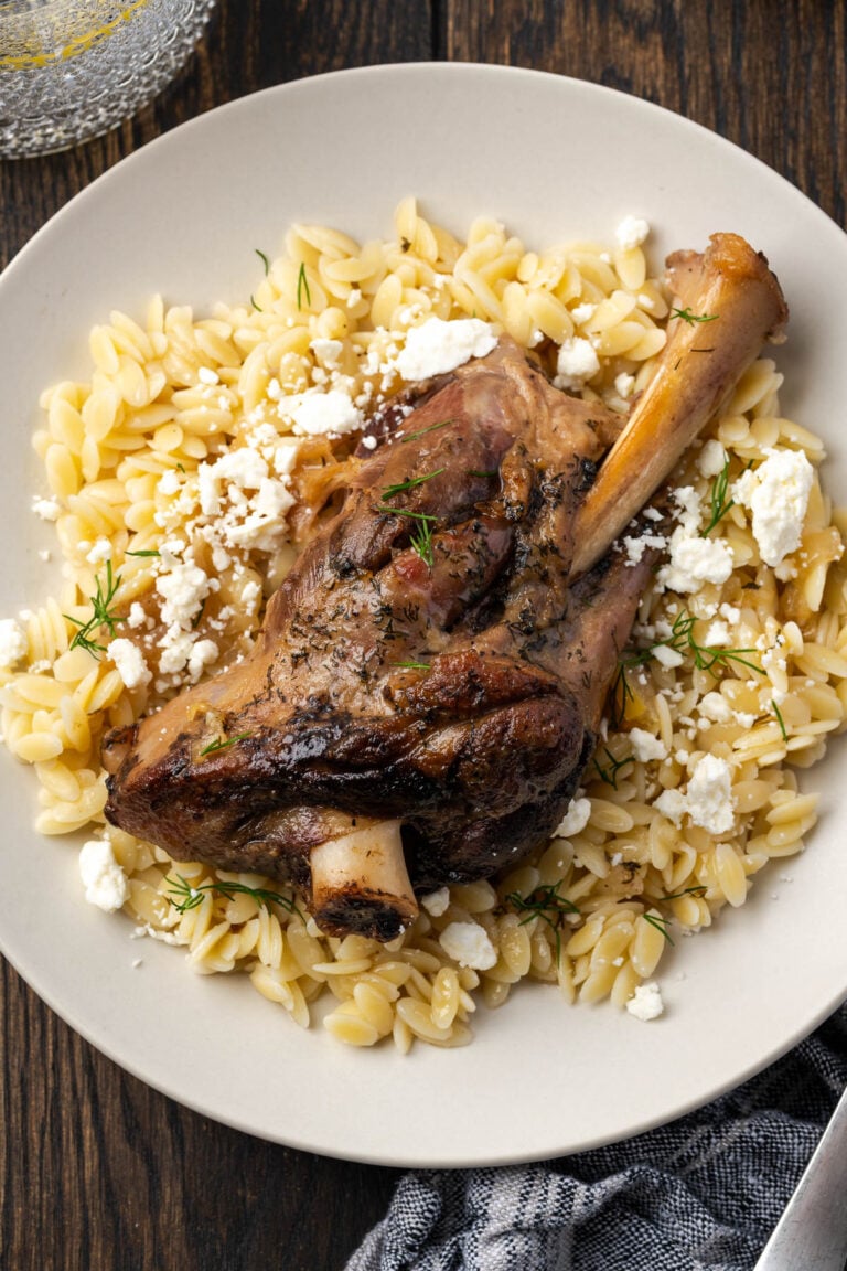 A plate with a roasted lamb shank served over orzo pasta, garnished with crumbled feta cheese and fresh herbs, on a wooden table.