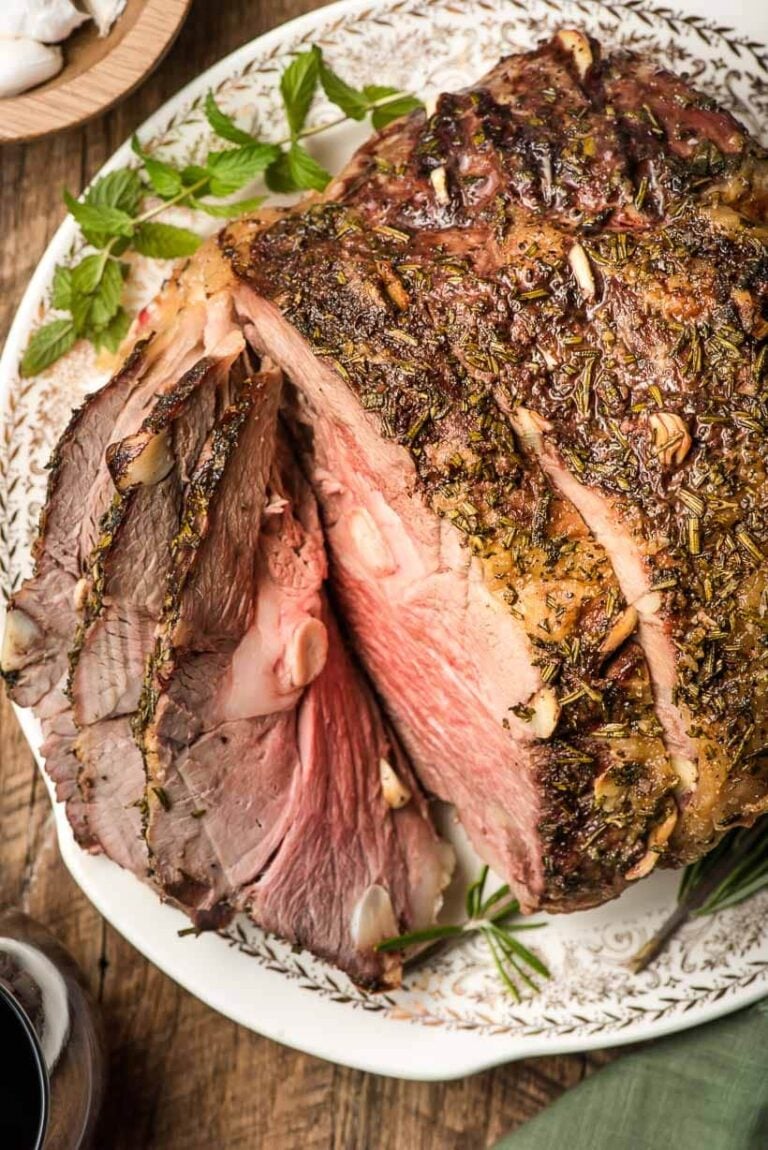 A close-up of a roast leg of lamb, sliced to show juicy, pink meat inside. The roast is seasoned with herbs and garlic and served on a white patterned plate, garnished with fresh herbs.