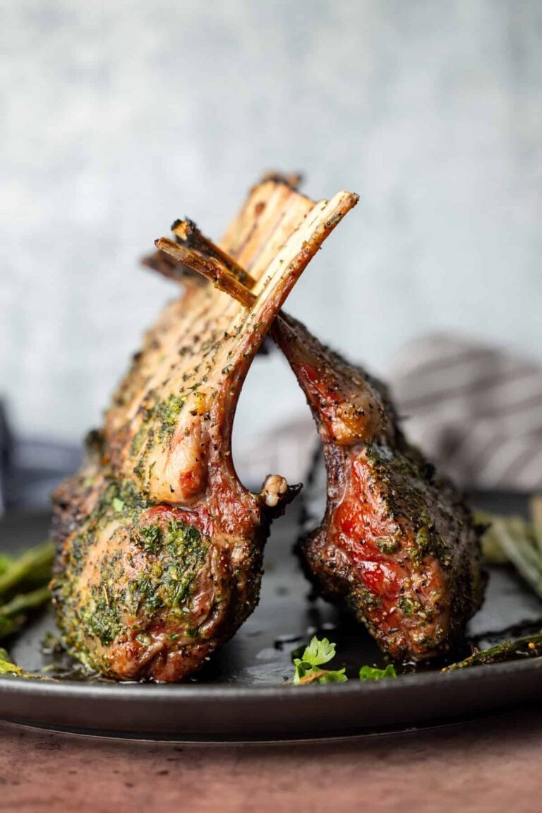 Two herb-crusted racks of lamb are arranged upright on a dark plate, their bones crossed. The meat is browned and garnished with herbs, with a blurred background and a sprig of parsley on the plate.