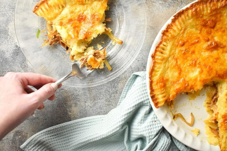A hand holding a fork takes a bite of savory pie filled with shredded meat and vegetables. Part of the pie is missing, revealing its filling. The pie sits on a plate next to a blue textured cloth.