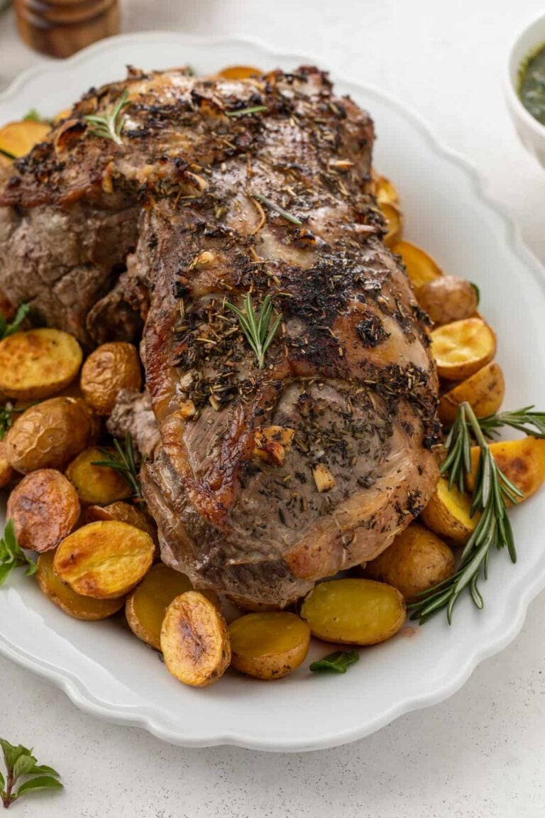 A roasted boneless leg of lamb garnished with herbs sits on a white platter, surrounded by golden roasted baby potatoes and sprigs of fresh rosemary.