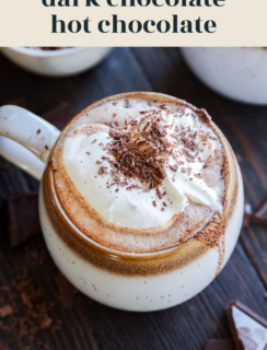 A mug of dark chocolate hot chocolate topped with whipped cream and chocolate shavings sits on a wooden table. Pieces of dark chocolate are scattered nearby. The text above reads, dark chocolate hot chocolate.