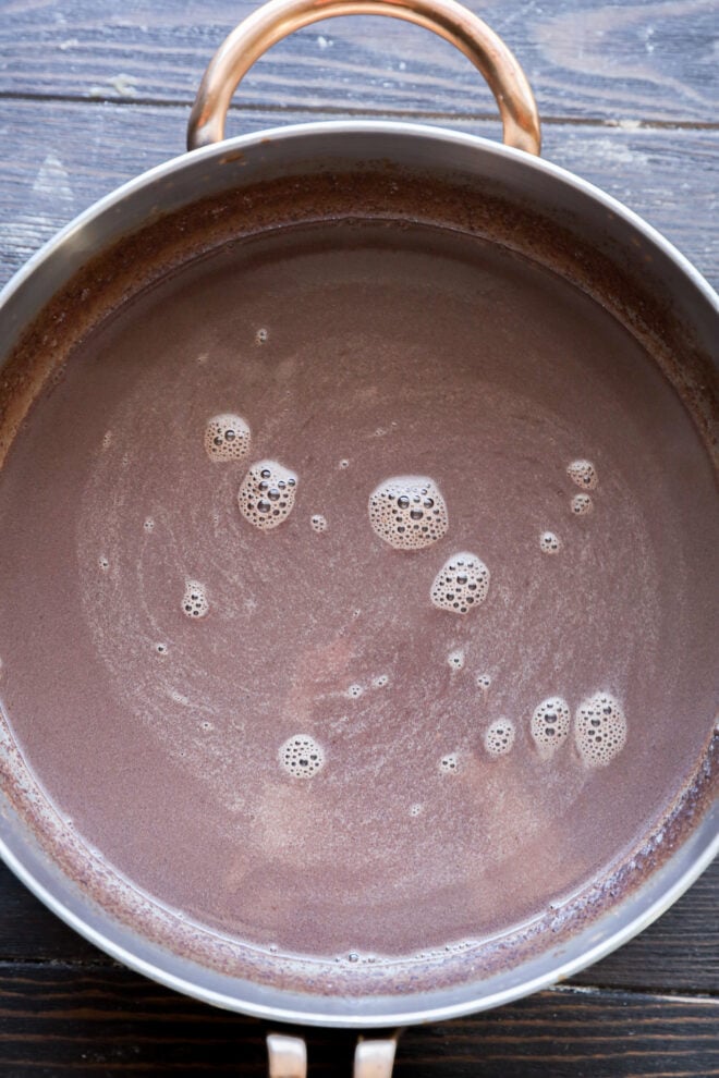 A metal pot filled with creamy hot chocolate, with small bubbles and foam on the surface, sits on a dark wooden table.