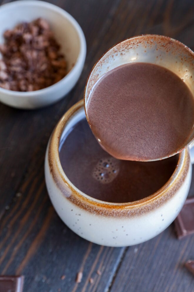A ladle pours creamy hot chocolate into a speckled ceramic mug on a wooden surface, with a bowl of cocoa powder and chocolate pieces nearby.