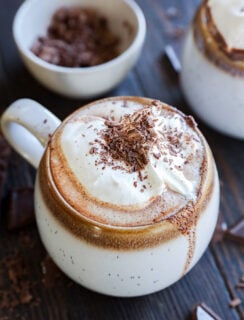 A mug of hot chocolate topped with whipped cream and chocolate shavings sits on a dark wooden surface; a bowl of chocolate shavings and another mug are in the background.