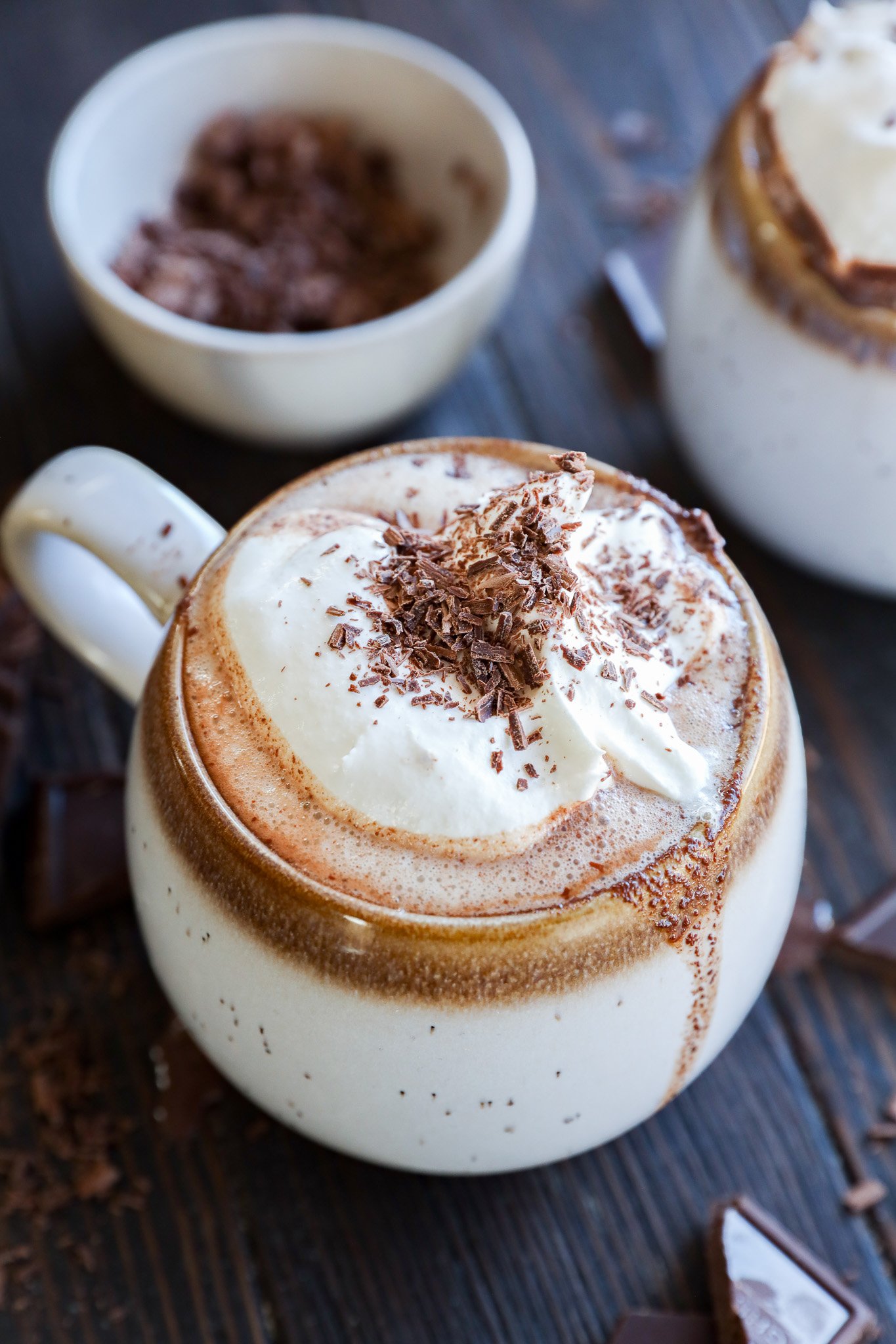 A mug of hot chocolate topped with whipped cream and chocolate shavings sits on a dark wooden surface; a bowl of chocolate shavings and another mug are in the background.