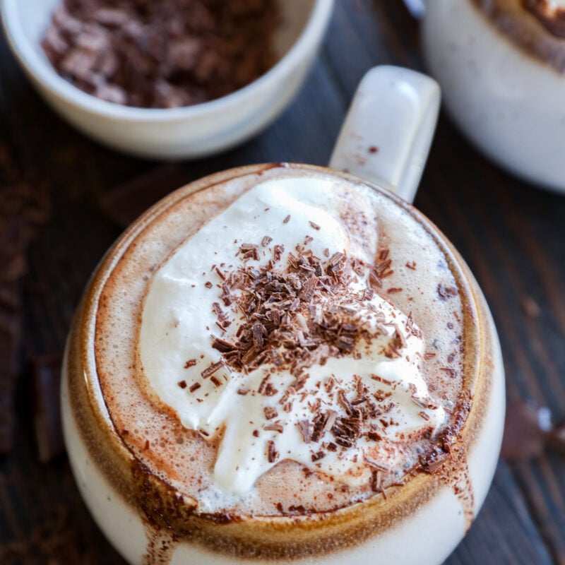 A mug of hot chocolate topped with whipped cream and chocolate shavings sits on a wooden table, with a bowl of grated chocolate in the background.