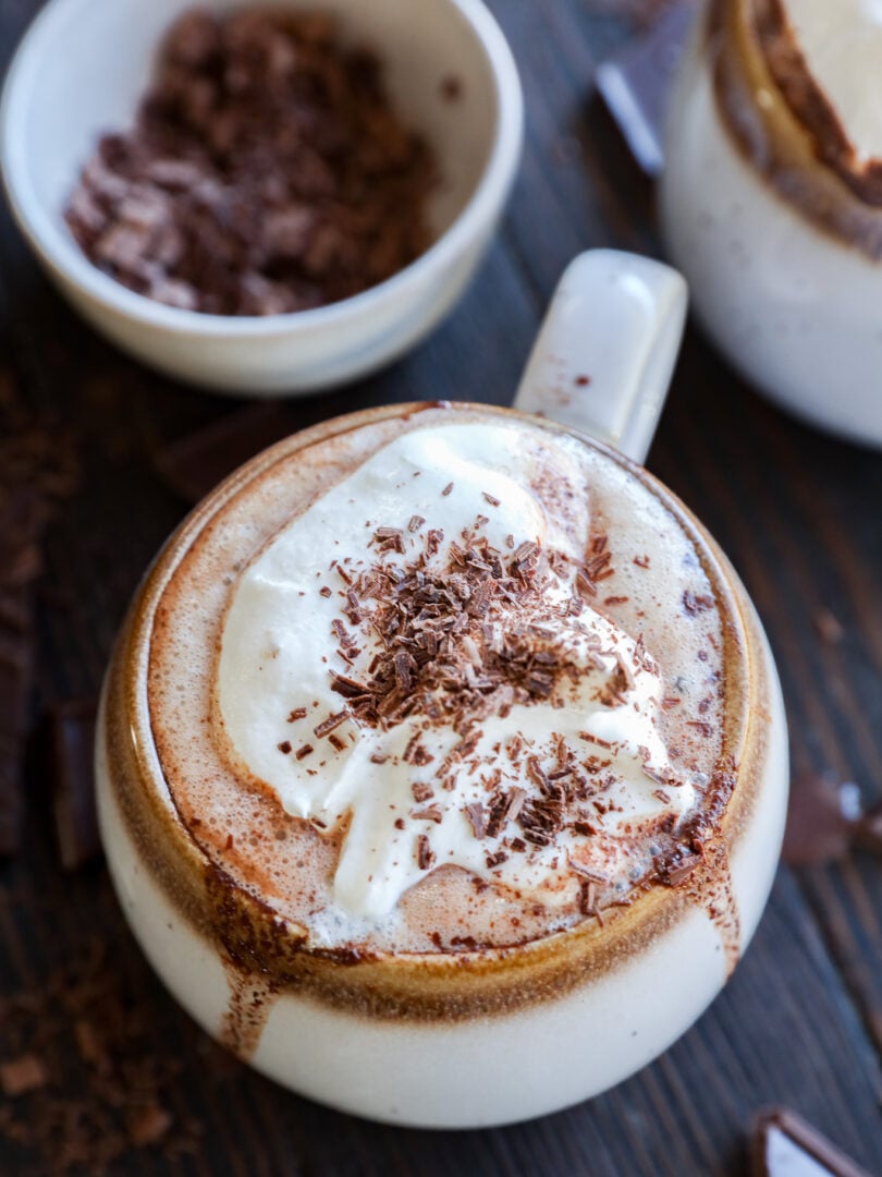 A mug of hot chocolate topped with whipped cream and chocolate shavings sits on a wooden table, with a bowl of grated chocolate in the background.