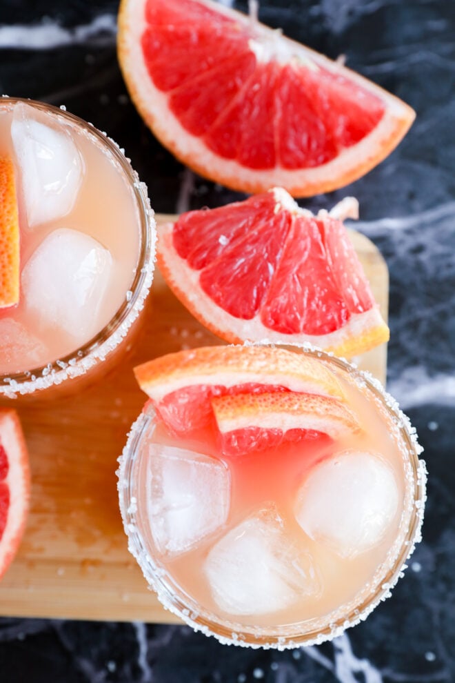 Two glasses of Grapefruit Vodka Cocktail with ice and salted rims, garnished with grapefruit slices. Fresh grapefruit wedges are placed nearby on a dark marble surface and a wooden board.