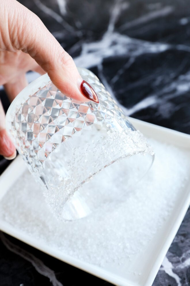 A hand dips the rim of a textured glass into coarse white salt. The background is a dark, marbled surface.
