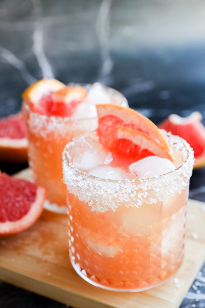 Two glasses of Grapefruit Vodka Cocktail with ice, salt-rimmed edges, and grapefruit wedges as garnish sit on a wooden board, with grapefruit slices in the background.