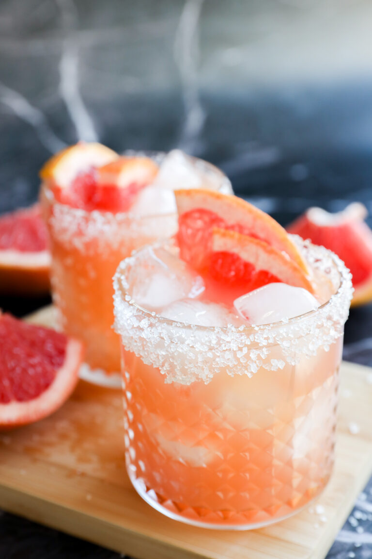 Two glasses of Grapefruit Vodka Cocktail with ice, salt-rimmed edges, and grapefruit wedges as garnish sit on a wooden board, with grapefruit slices in the background.