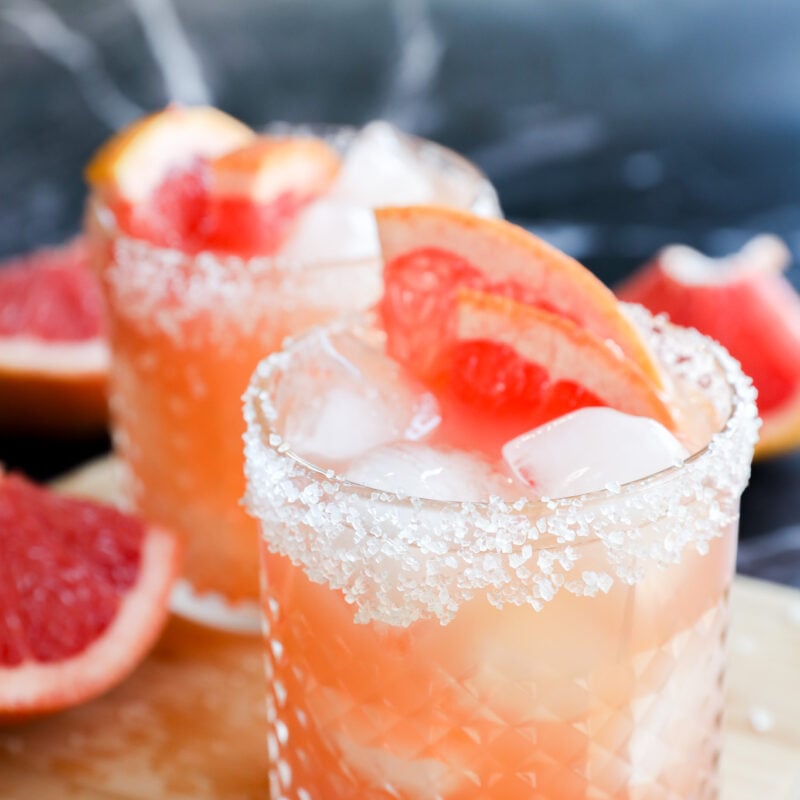 Two glasses of Grapefruit Vodka Cocktail with ice, salt-rimmed edges, and grapefruit wedges as garnish sit on a wooden board, with grapefruit slices in the background.