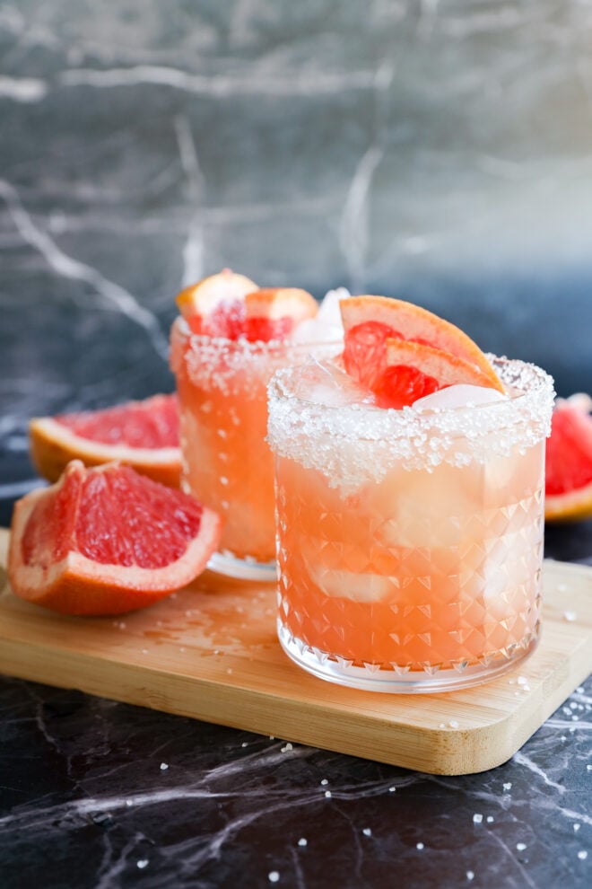 Two glasses of Grapefruit Vodka Cocktail with ice and salted rims sit on a wooden board, garnished with grapefruit wedges. Fresh grapefruit halves are nearby, all set against a marble background.