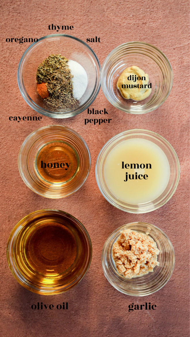 Overhead view of seven glass bowls on a brown surface, each labeled with ingredients for Greek Lemon Chicken and Potatoes: oregano, thyme, salt, cayenne, black pepper, Dijon mustard, honey, lemon juice, olive oil, and garlic.