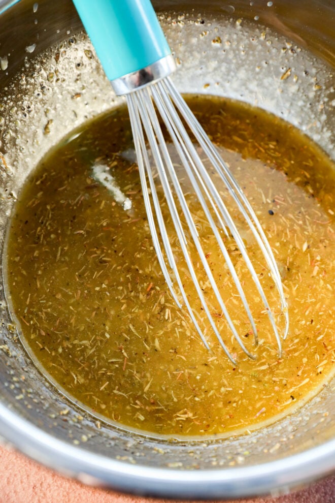 A metal bowl containing a yellow-brown herb marinade with a whisk that has a turquoise handle.