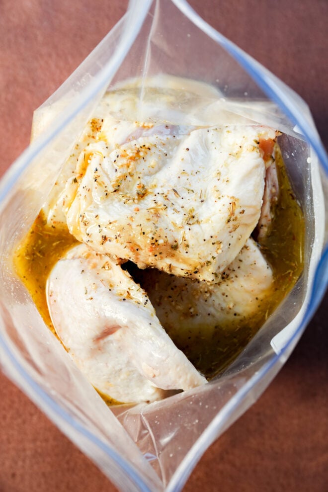 Raw chicken pieces marinating in a clear plastic zip-top bag with an herb marinade