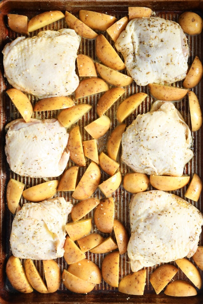 Raw chicken thighs and seasoned potato wedges are arranged on a baking sheet