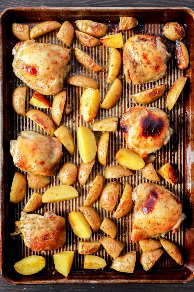 A baking sheet with six Greek Lemon Chicken and Potatoes—roasted chicken thighs and golden-brown potato wedges, all seasoned and cooked, arranged evenly on a lined pan.
