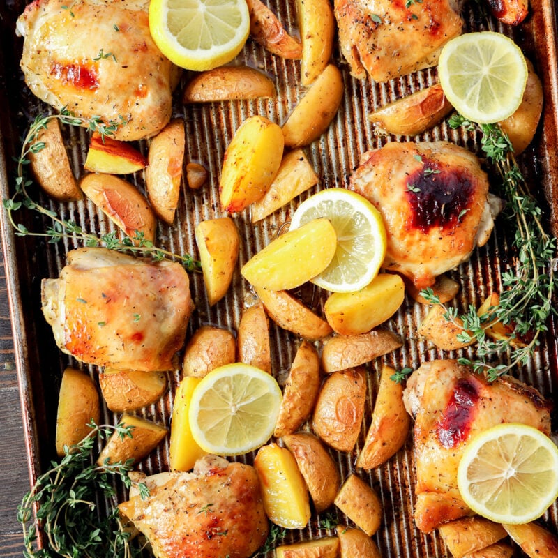 A baking sheet with Greek Lemon Chicken and Potatoes, golden wedges, lemon slices, and fresh thyme sprigs, all arranged on a striped parchment lining. The food appears well-cooked and beautifully garnished for serving.